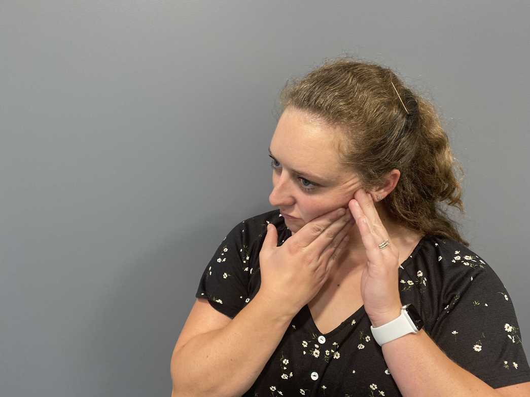 Women holding side of her face and jaw with 2 hands wearing black shirt with flowers and grey wall in background