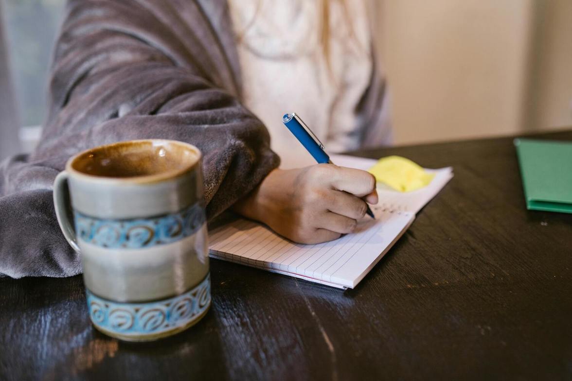 A person wrapped in a cozy blanket, writing in a notebook at a wooden table with a mug beside them—capturing a quiet moment of reflection and gratitude.