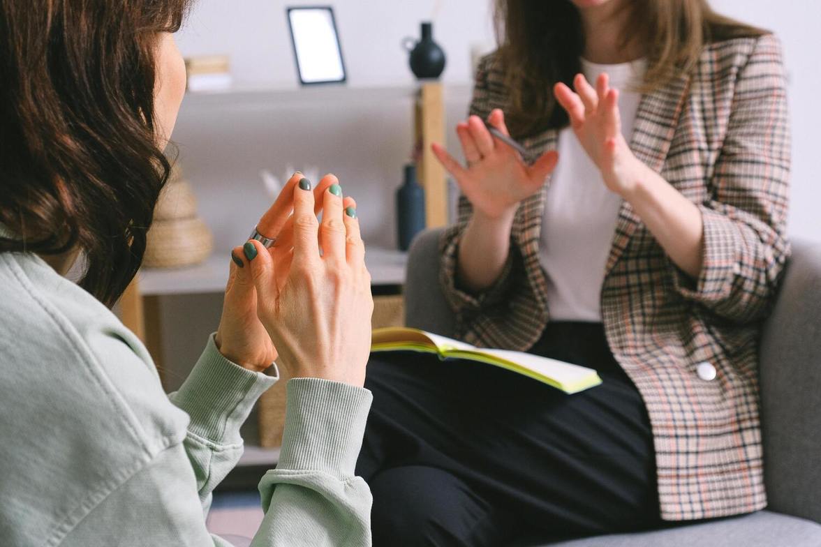 Two women sitting across from each other during an individual therapy session, therapist holding pen with notebook on lap, supportive and relaxed conversation