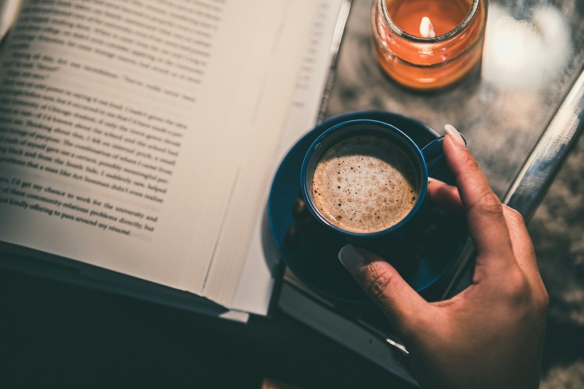 Close-up of a hand holding a warm coffee beside an open book and candle, suggesting a relaxing moment.