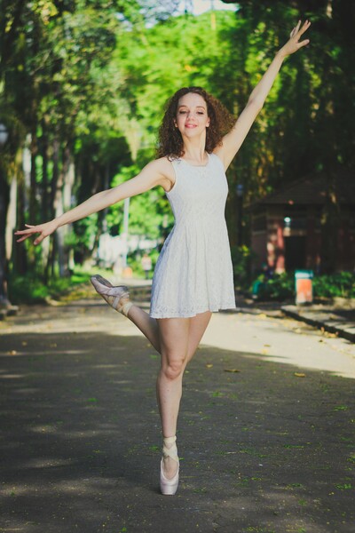 A ballerina is standing in the middle of a tree lined street, she is up on the toes of one foot, en pointe, with her arms in the air