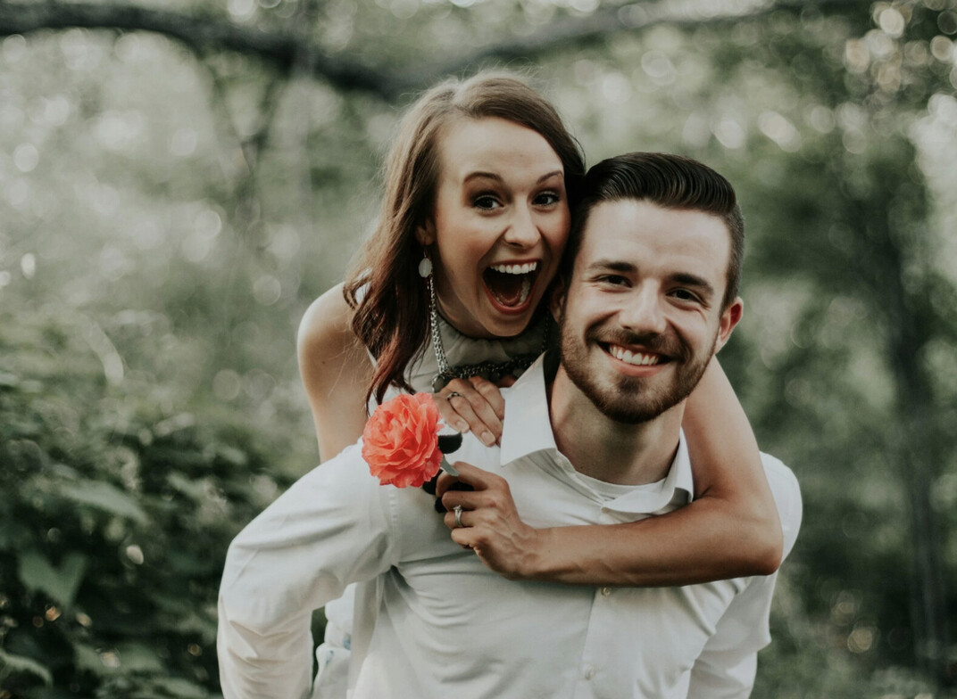 Female with brown hair and orange flower on the back of her boyfriend. Both people are very excited and smiling. 