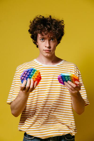 A white young male with wearing yellow and white stripped shirt, holding a rainbow colour fidget toy with a yellow solid colour background