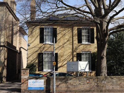 Two‑storey, yellow‑brick building with black shutters, partially shaded by a large tree in front. A low brick wall runs along the front, a tall signboard above displays the name ‘The Beechwood Practice.’