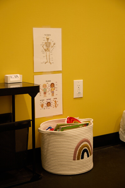 Rainbow on bucket holding children toys with black stand beside and Nova business cards on top and laminated posters on the yellow wall