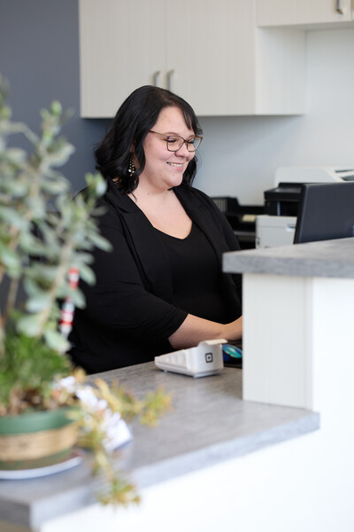 Woman sitting at front clinic reception desk wearing black jacket and green plant on desk