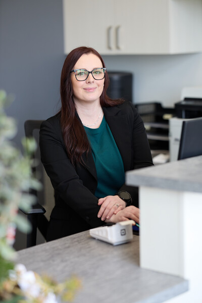 Woman sitting behind reception desk wearing black jacket with computer and plant on desk