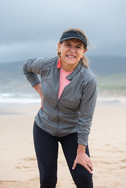 A older woman on the beach holds her back and leg from pain in both legs from sciatica