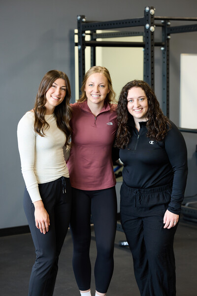 Three women standing in a clinic gym wearing a white t shirt, pink sweater and black sweater