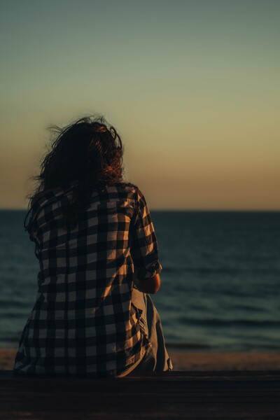 Shows back of woman with long brown hair sitting on beach looking out over lake towards horizon.