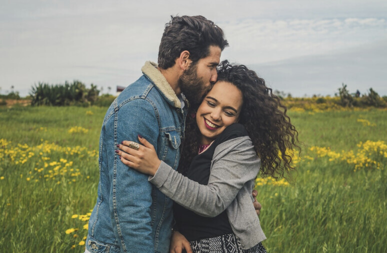 Cis-male with brown hair in a jean jacket kissing a woman with black curly hair. The couple is outside and look happy. 