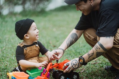 An Indigenous man with tattoos reveling sadness, plays warmly with his son who smiles broadly at him. 