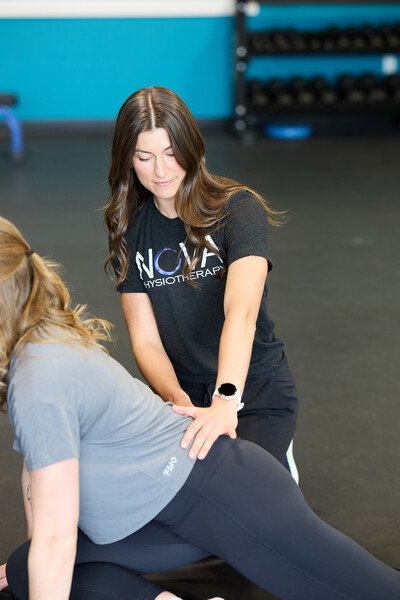 One woman who is a physiotherapist wearing a Nova shirt helping another women stretch her hips in a clinic gym