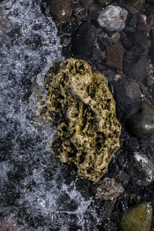 Water flowing forcefully against a rough rock in a shallow stream, symbolizing structural resistance, pressure, and the cumulative impact of systemic burnout.