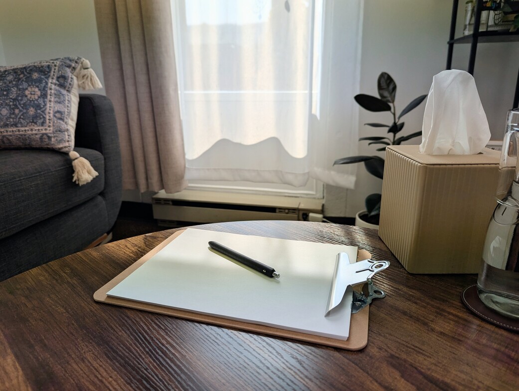 A clipboard with paper and a pen are on a coffee table in Alicia Norman's counselling office. Sunlight streams in through the window.