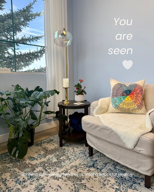 A bright, welcoming therapy room in St. Albert. A grey armchair with a white faux fur throw and an 'emotions wheel' pillow sits next to a large window with natural light, a monstera plant, and a small side table with a candle.
